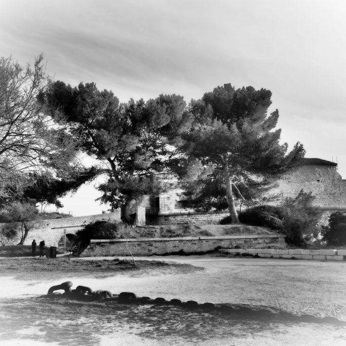 Paysage en noir et blanc avec des arbres, un bâtiment et des personnes au bord de l'eau.