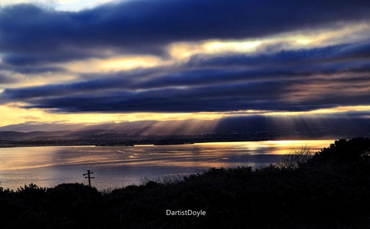 Coucher de soleil sur un lac, avec des nuages sombres et des rayons de lumière.